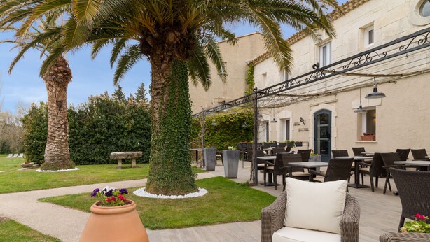 An outdoor seating area with wicker furniture, a large planter with flowers, and a palm tree. - Val Baussenc, The Originals Relais (Maussane-les-Alpilles)