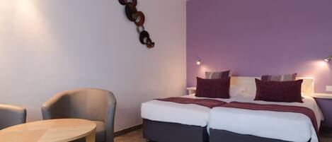 A modern hotel room with a bed, two bedside tables, a small round table, and a wall-mounted light fixture.