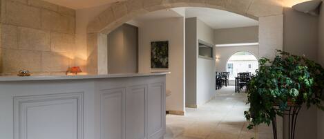 A reception area with a stone archway, a reception desk, a potted plant, and a dining area with tables and chairs in the background.