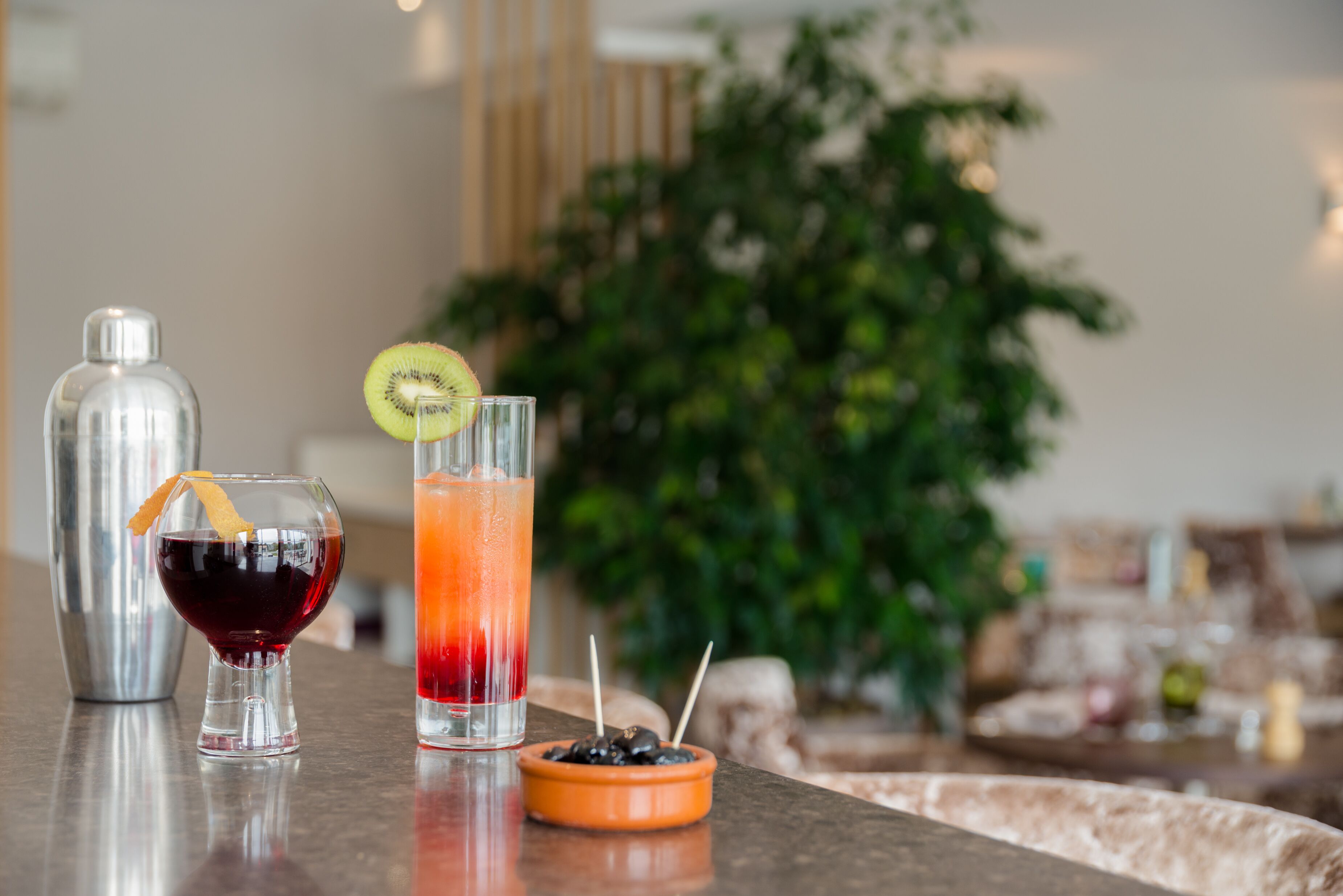 Zona de bar con dos bebidas, un shaker de cócteles y un cuenco pequeño de aceitunas.