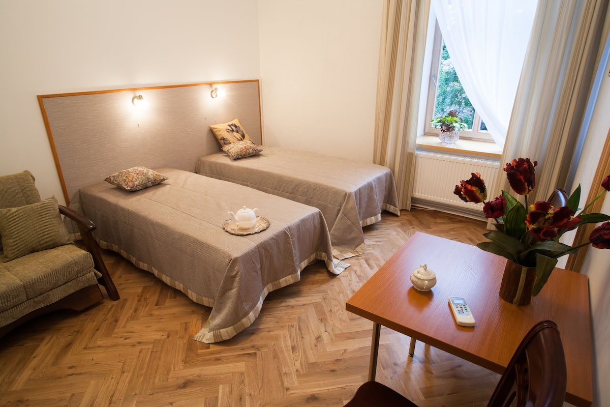 A hotel room with two single beds, a wooden floor, a small table with a vase of flowers, and a window with curtains.