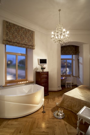 A bedroom with a large bathtub, a chandelier, a wooden floor, and a view of the outside through a window. - Florens Boutique (Vilnius)