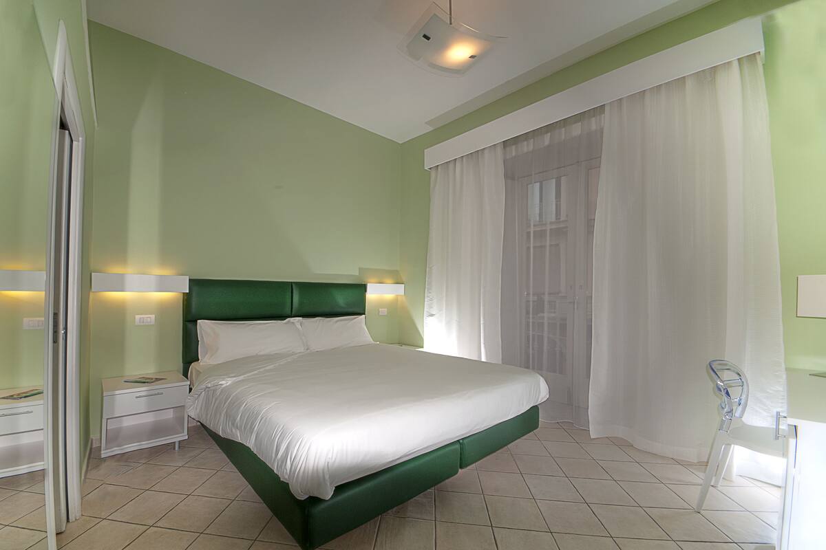 Une chambre à coucher bien rangée, avec un lit vert, du linge de lit blanc et une grande fenêtre ornée de rideaux légers.