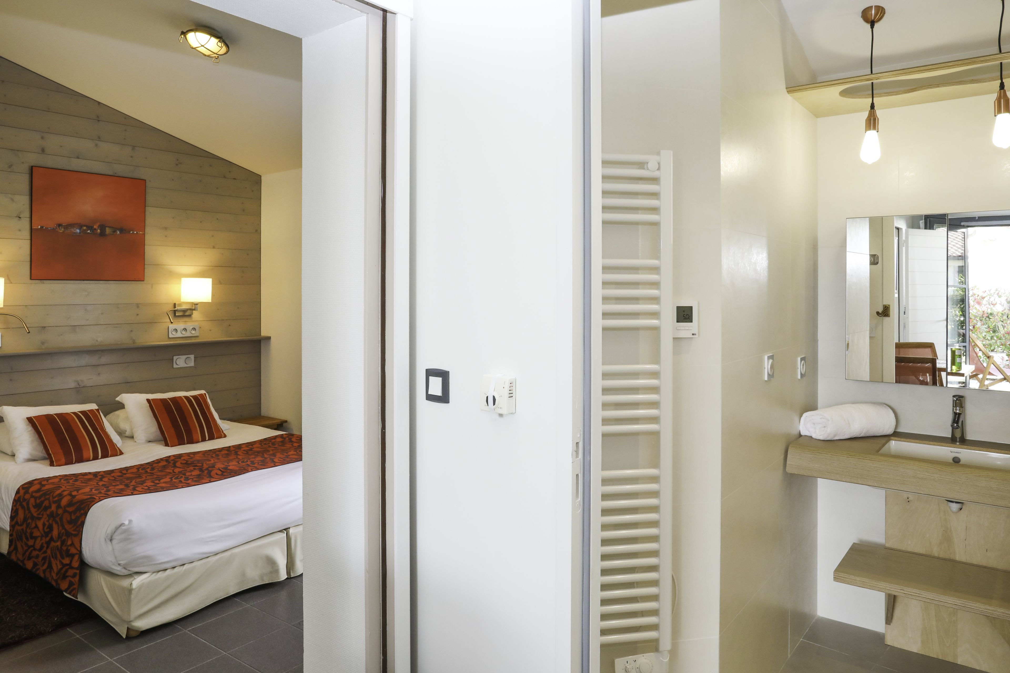 Une chambre d’hôtel avec un lit, des lampes de chevet, une tête de lit en bois, un grand miroir, une coiffeuse en bois et une fenêtre donnant sur un paysage.