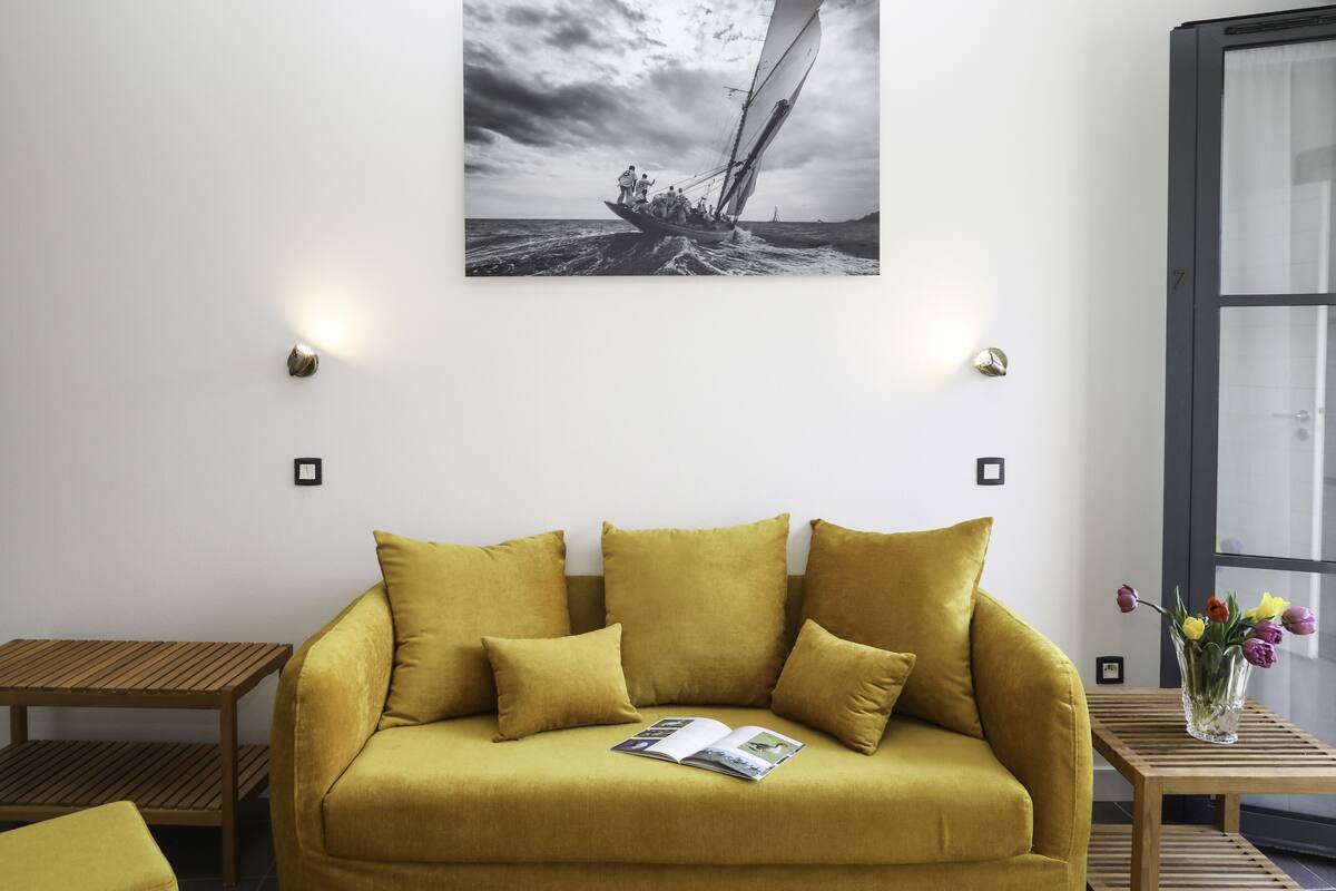 Una habitación con un sofá color mostaza, una mesita de madera, una fotografía enmarcada en la pared y una ventana con vistas al mar.