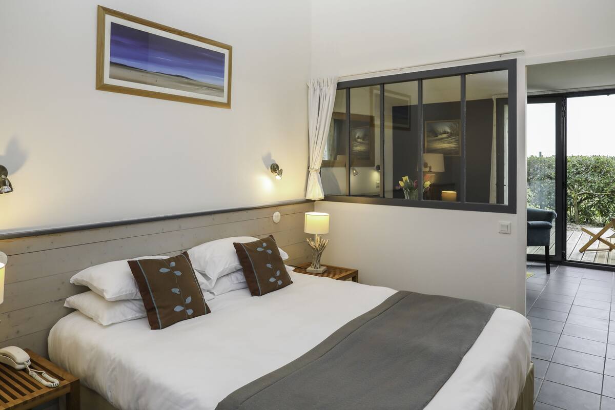 Una habitación de hotel con cama, lámpara de noche, teléfono, ventana con cortinas y un cuadro en la pared.