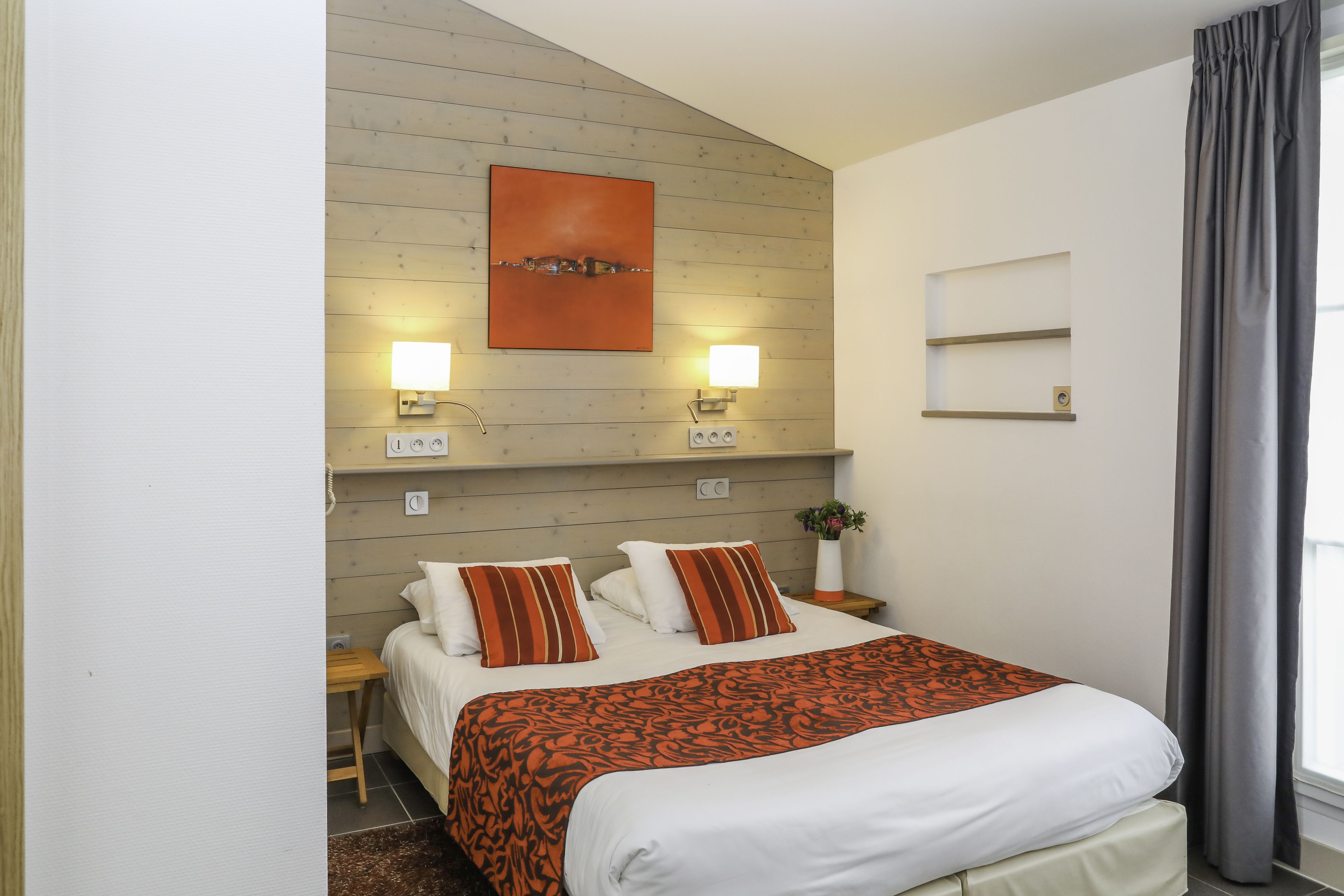 Ein modernes Hotelzimmer mit einem Holz-Kopfteil, einem großen Bett mit weißer und orangefarbener Bettwäsche, zwei Nachttischlampen und Blick auf die Stadt durch ein Fenster mit grauen Vorhängen.