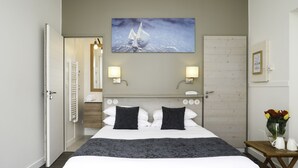 A hotel room with a bed, two bedside lamps, a painting of a sailboat, a wooden door, and a small table with a vase of flowers.