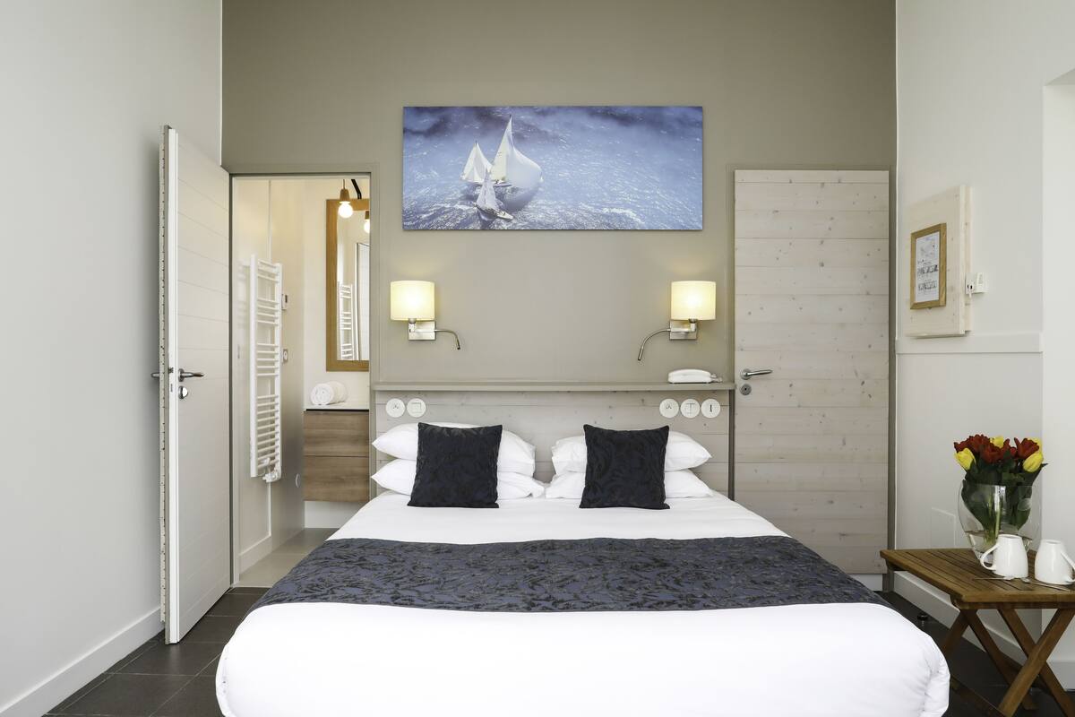 Una habitación de hotel con una cama, dos lámparas de noche, un cuadro de un velero, una puerta de madera y una pequeña mesa con un florero.