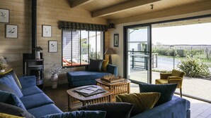 A cozy living room with a blue sofa, a wooden coffee table, and a chessboard. There is a fireplace, a large window, and a view of the outdoors.