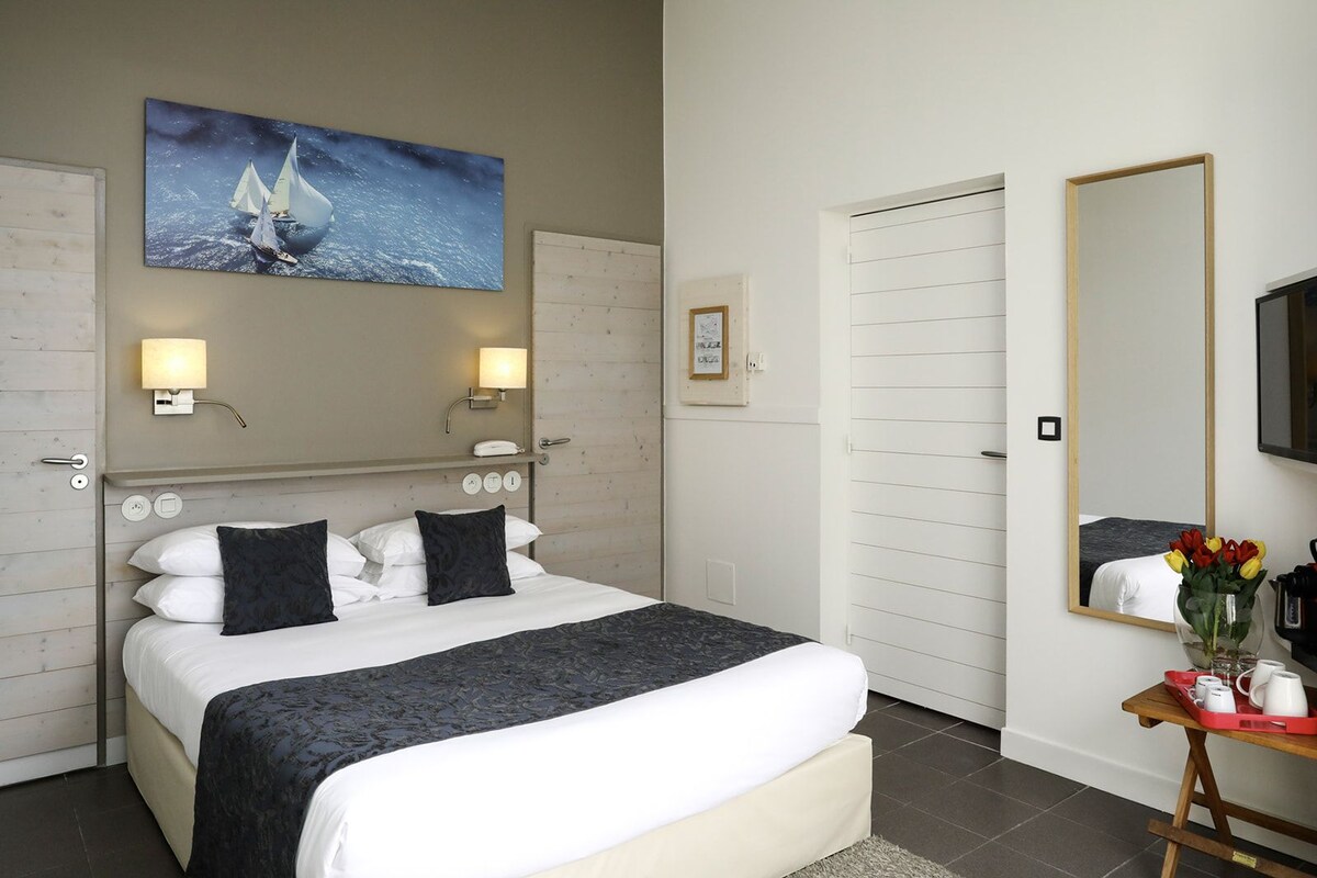 A hotel room with a bed, bedside lamps, a painting of sailboats, a mirror, and a small table with a vase of flowers.
