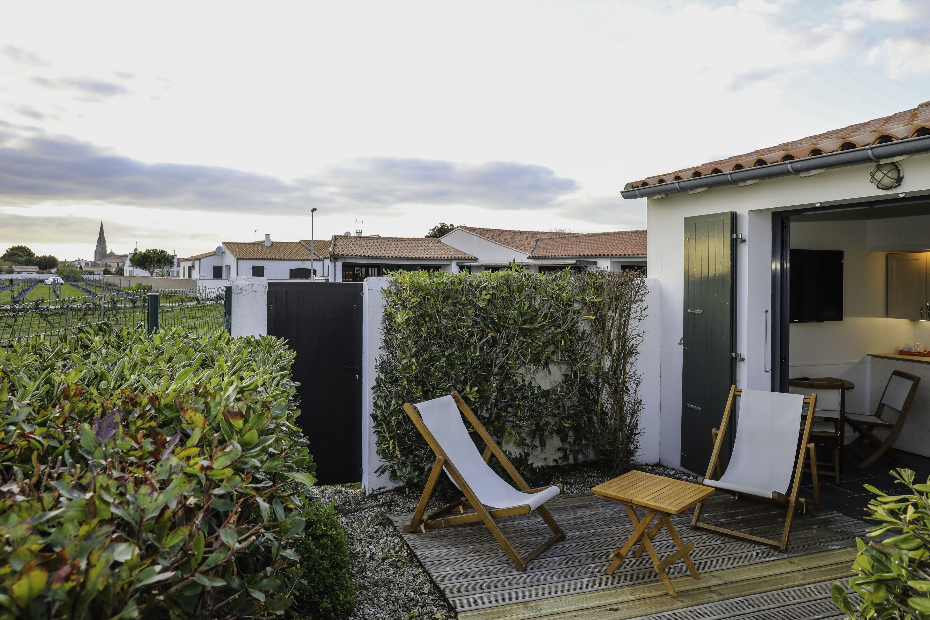 A patio area with two lounge chairs and a small table, surrounded by greenery and a white building with a tiled roof.