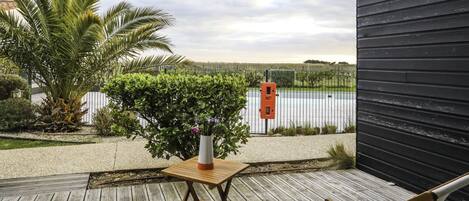 A wooden deck with two chairs and a small table, a potted plant, and a view of a body of water and a fence in the background.