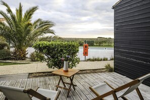 Una terraza de madera con dos sillas y una mesita, una planta en maceta, y vista a un cuerpo de agua y una cerca al fondo.