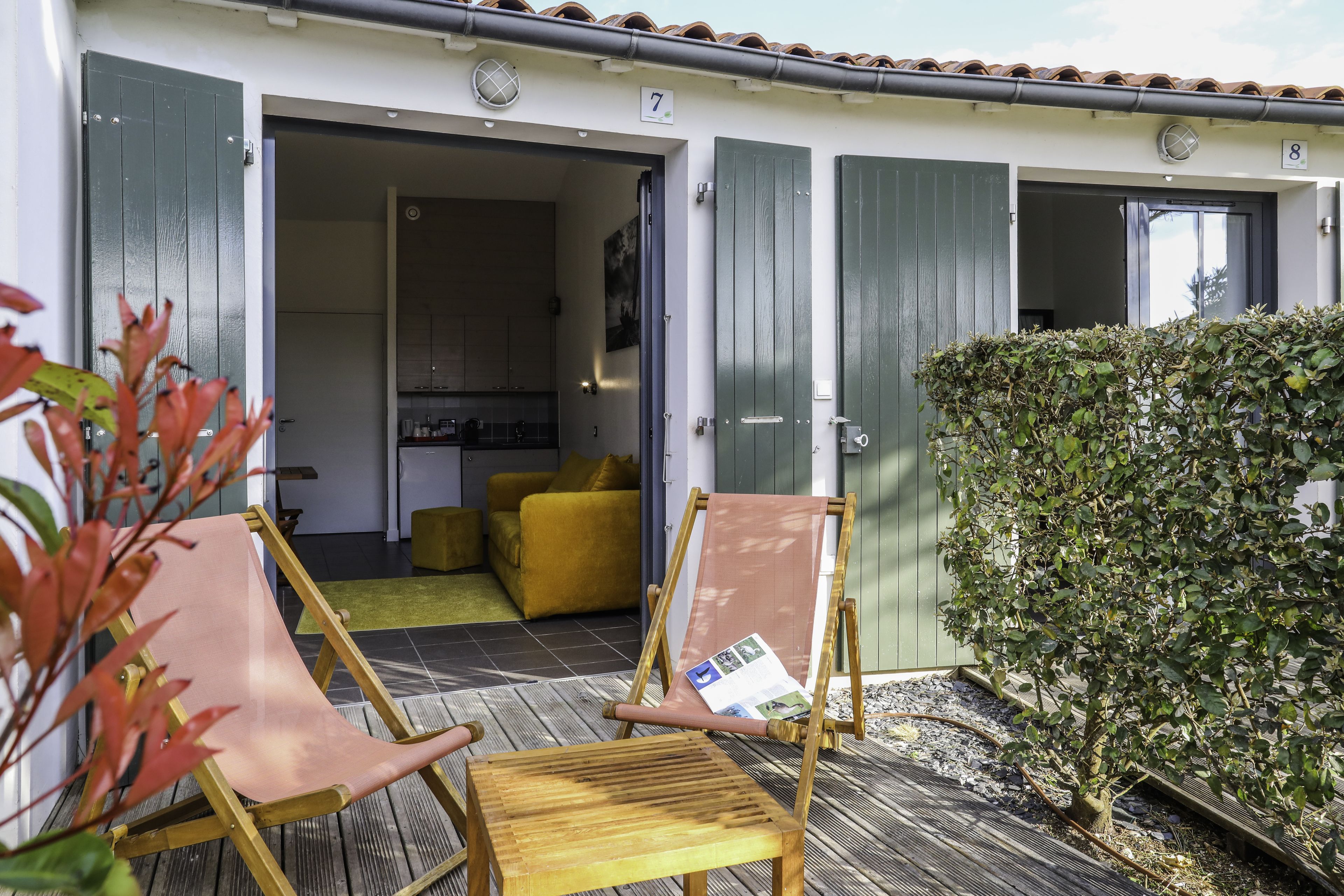 Quarto de hotel com varanda de madeira, sofá amarelo e sebe verde.