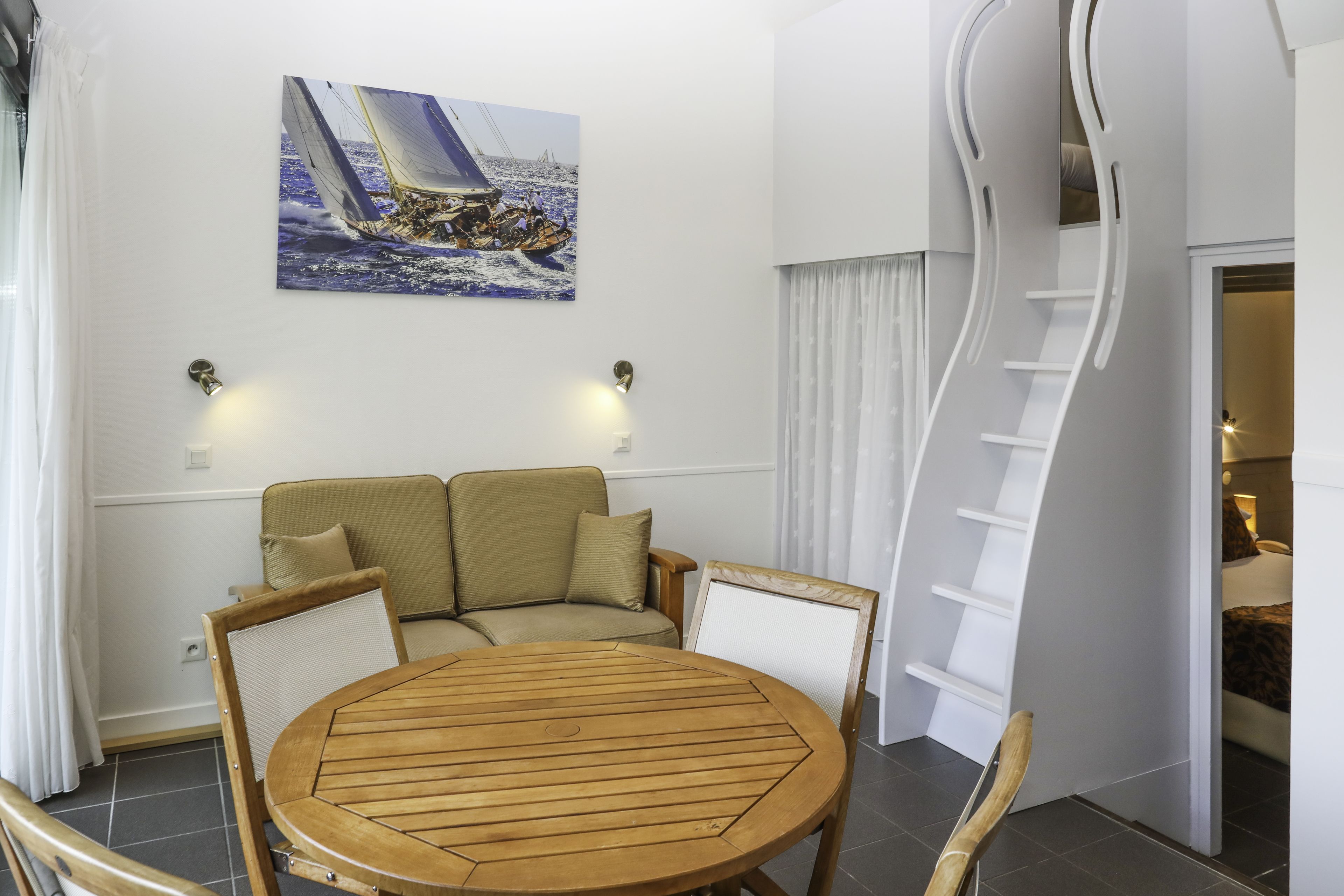 Une pièce comprenant une table ronde en bois, deux fauteuils rembourrés, un canapé, un tableau représentant un voilier et un escalier en colimaçon blanc.