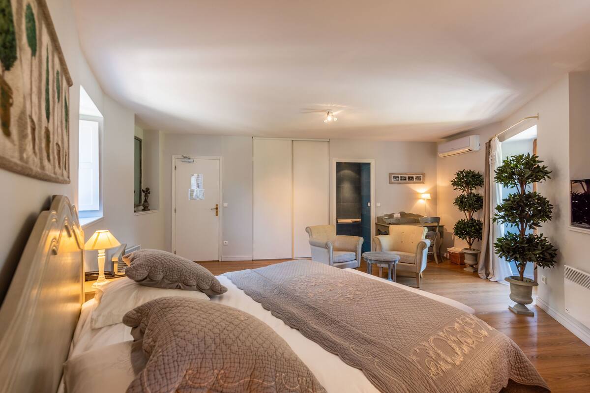 A bedroom with a bed, bedside lamps, a chair, a desk, and a potted plant.