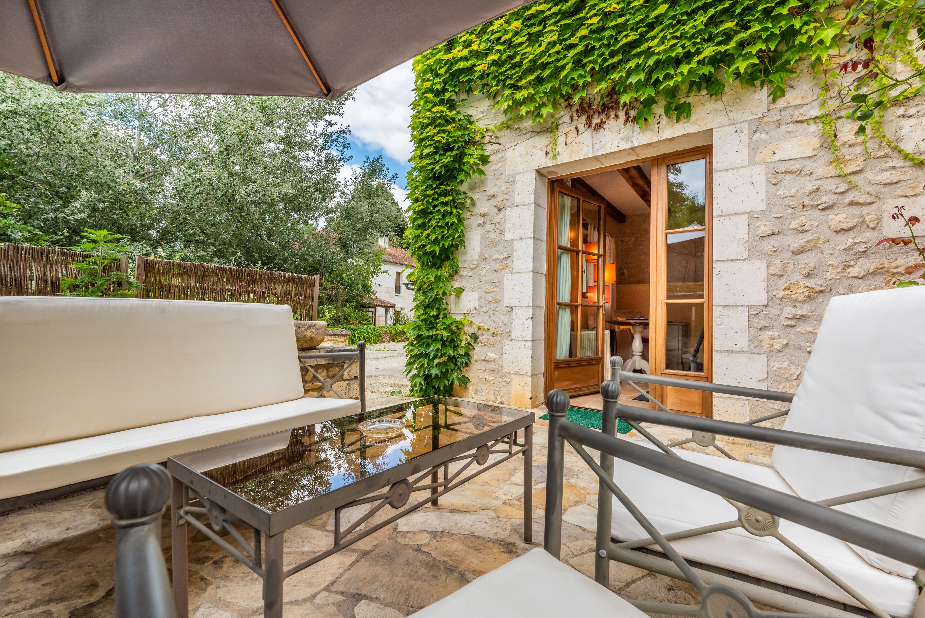 Un espace extérieur aménagé avec une table en pierre, des chaises blanches dotées de coussins et un mur en pierre agrémenté de verdure.