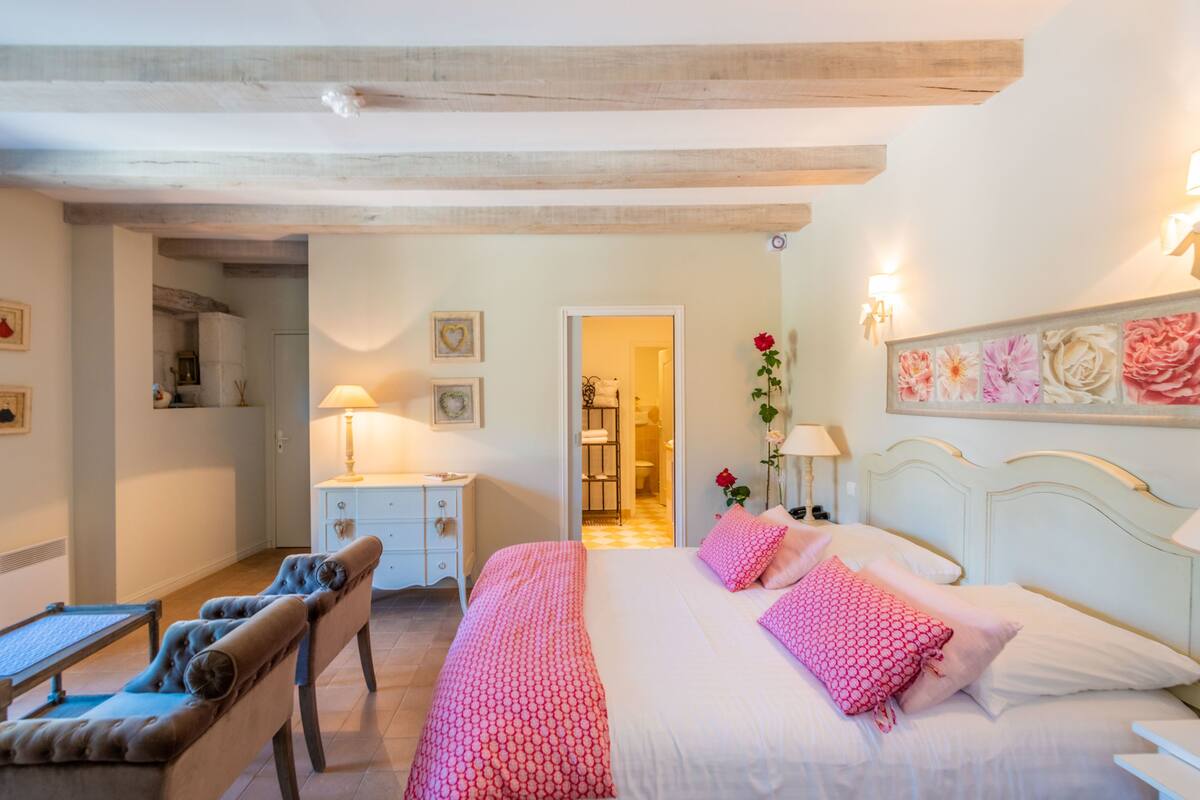 A bedroom with a bed, bedside table, lamp, chair, and a hallway leading to a bathroom.