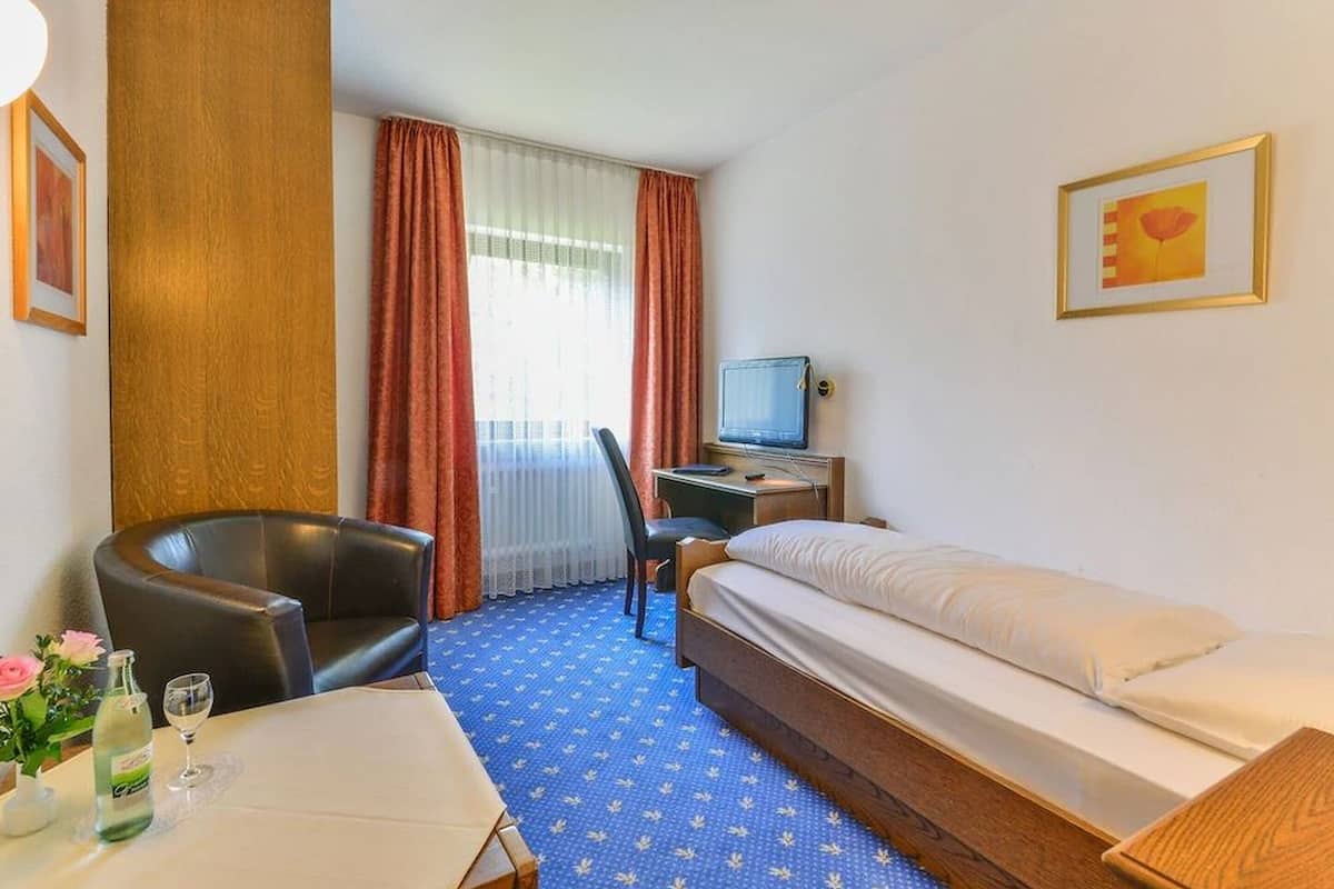 Una habitación de hotel con cama, escritorio con computadora, silla, mesa con un jarrón de flores y ventana con cortinas.