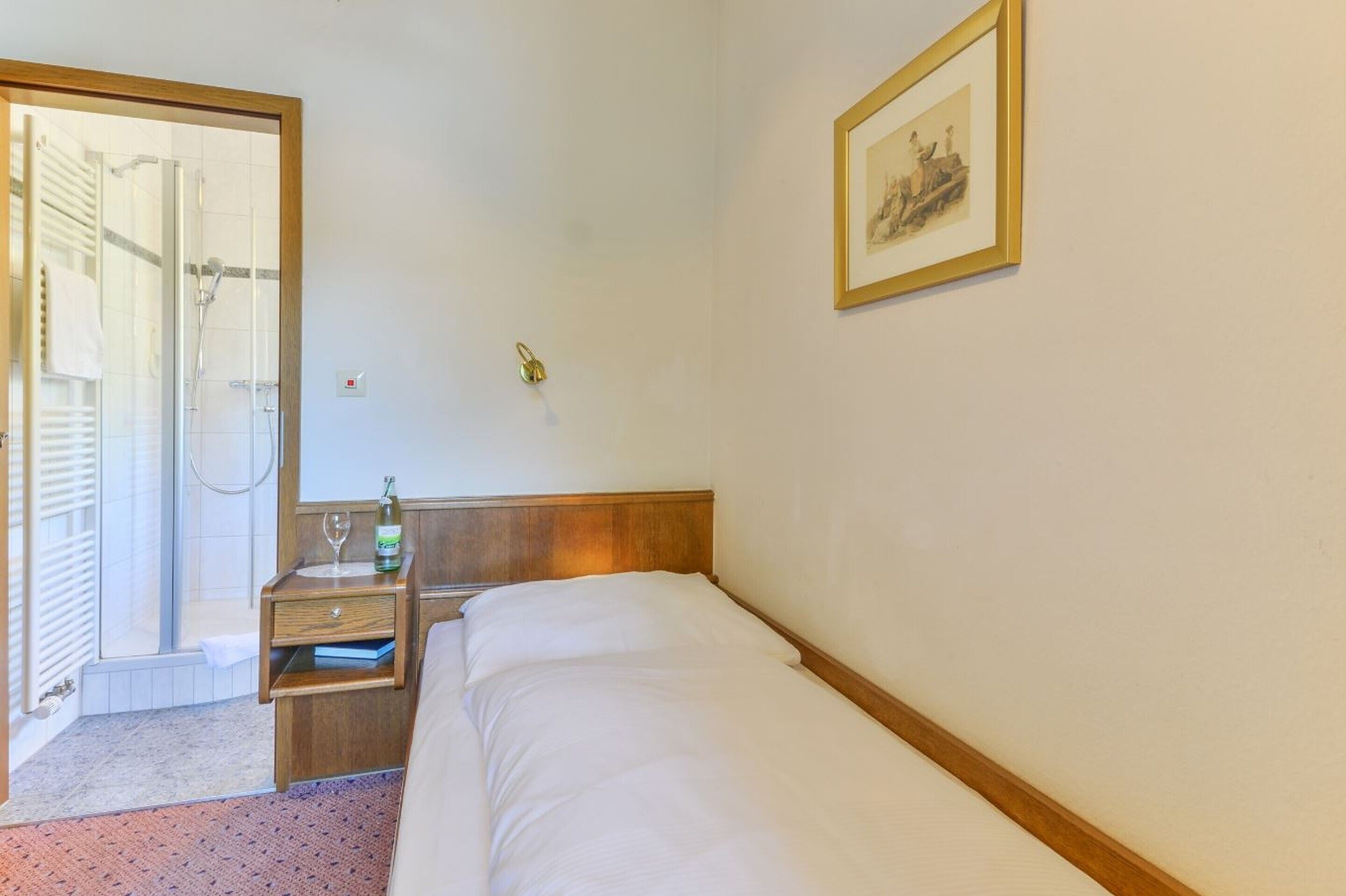 A hotel room with a bed, a bedside table with a bottle and glass, a framed picture on the wall, and a shower area visible through an open door.