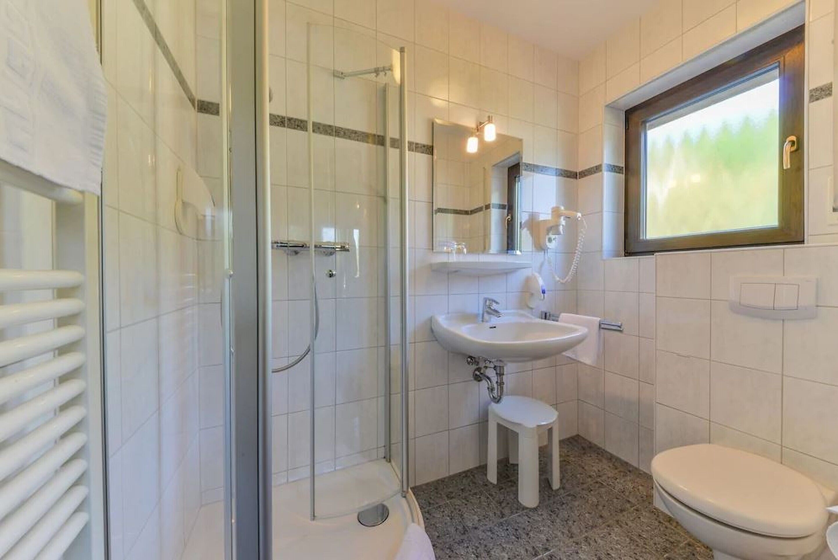 A modern bathroom with a glass-enclosed shower, a pedestal sink, a toilet, and a window with a view of greenery.