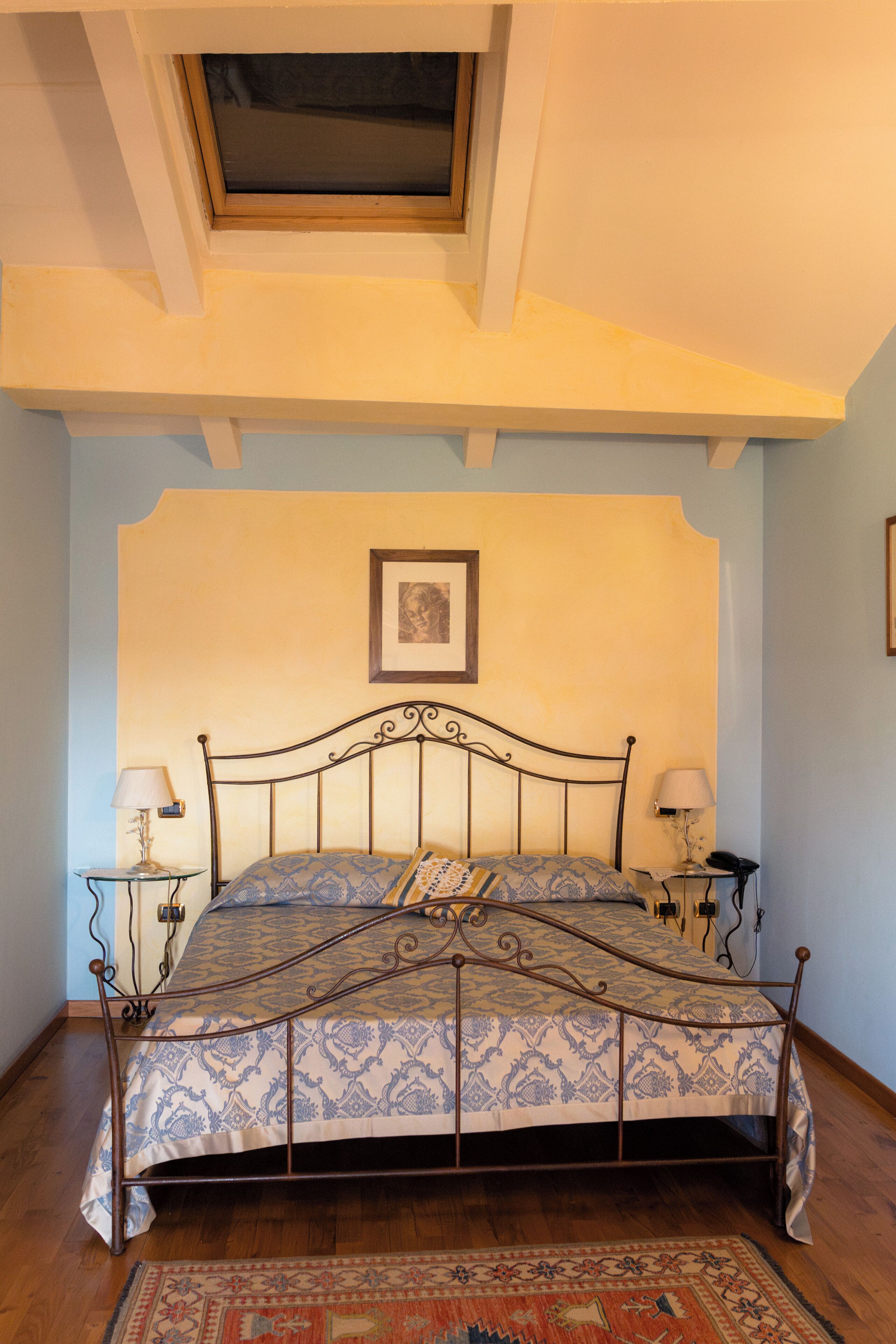 A bedroom with a metal bed frame, a patterned bedspread, two bedside lamps, a framed picture on the wall, and a skylight above the bed.