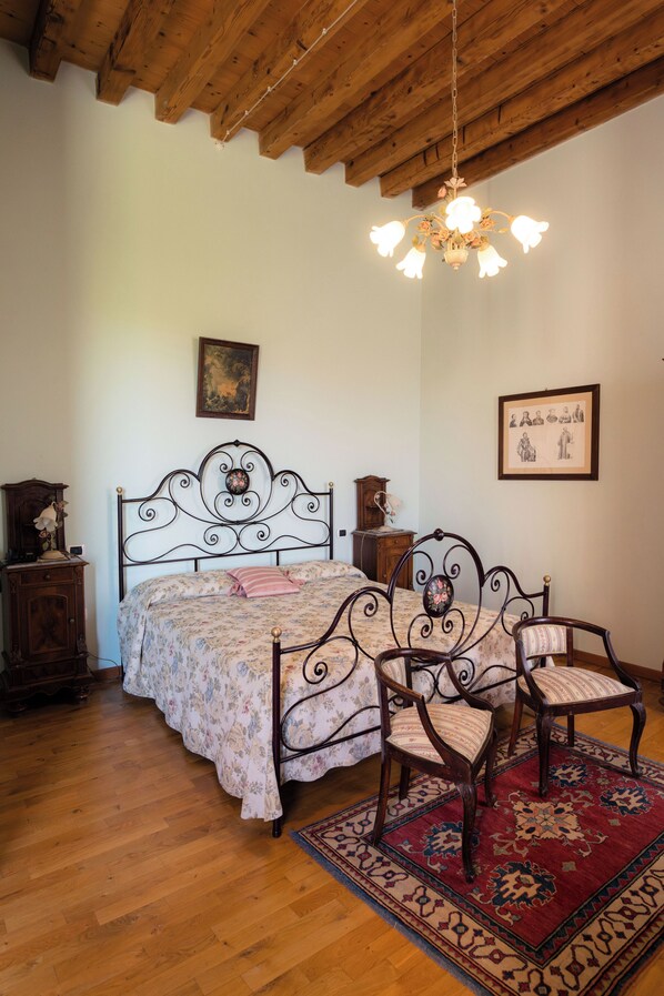 A bedroom with a wooden ceiling, a bed with a floral bedspread, two upholstered chairs, a red patterned rug, and a chandelier. - Villa Bongiovanni (San Bonifacio)