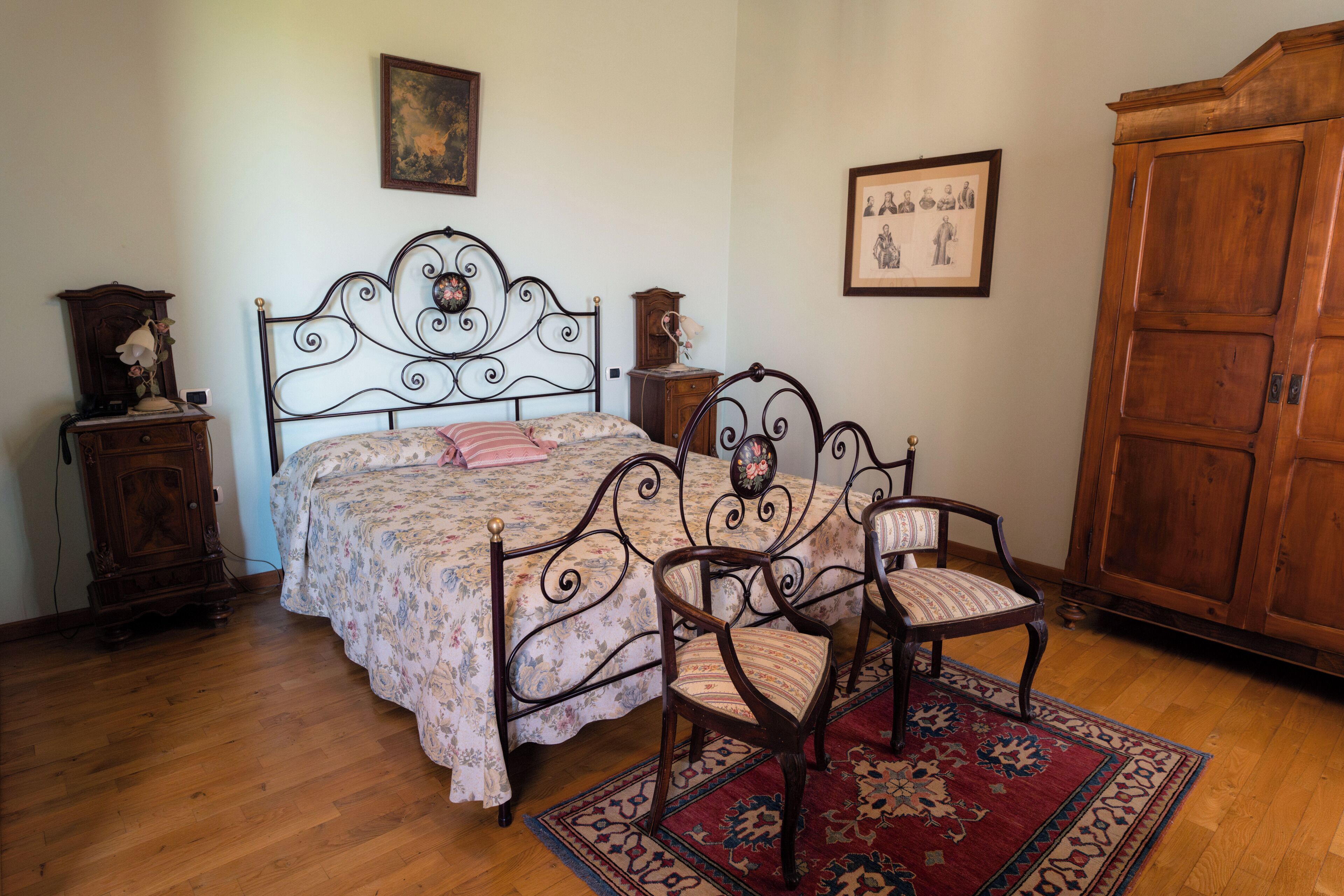A bedroom with a bed, two chairs, a wooden wardrobe, and a nightstand with a lamp.