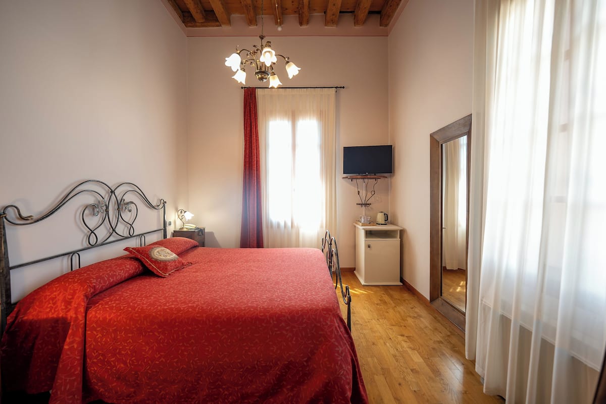 A bedroom with a wooden ceiling, a bed with a red bedspread, a bedside table with a lamp, a TV on a stand, and a window with curtains.