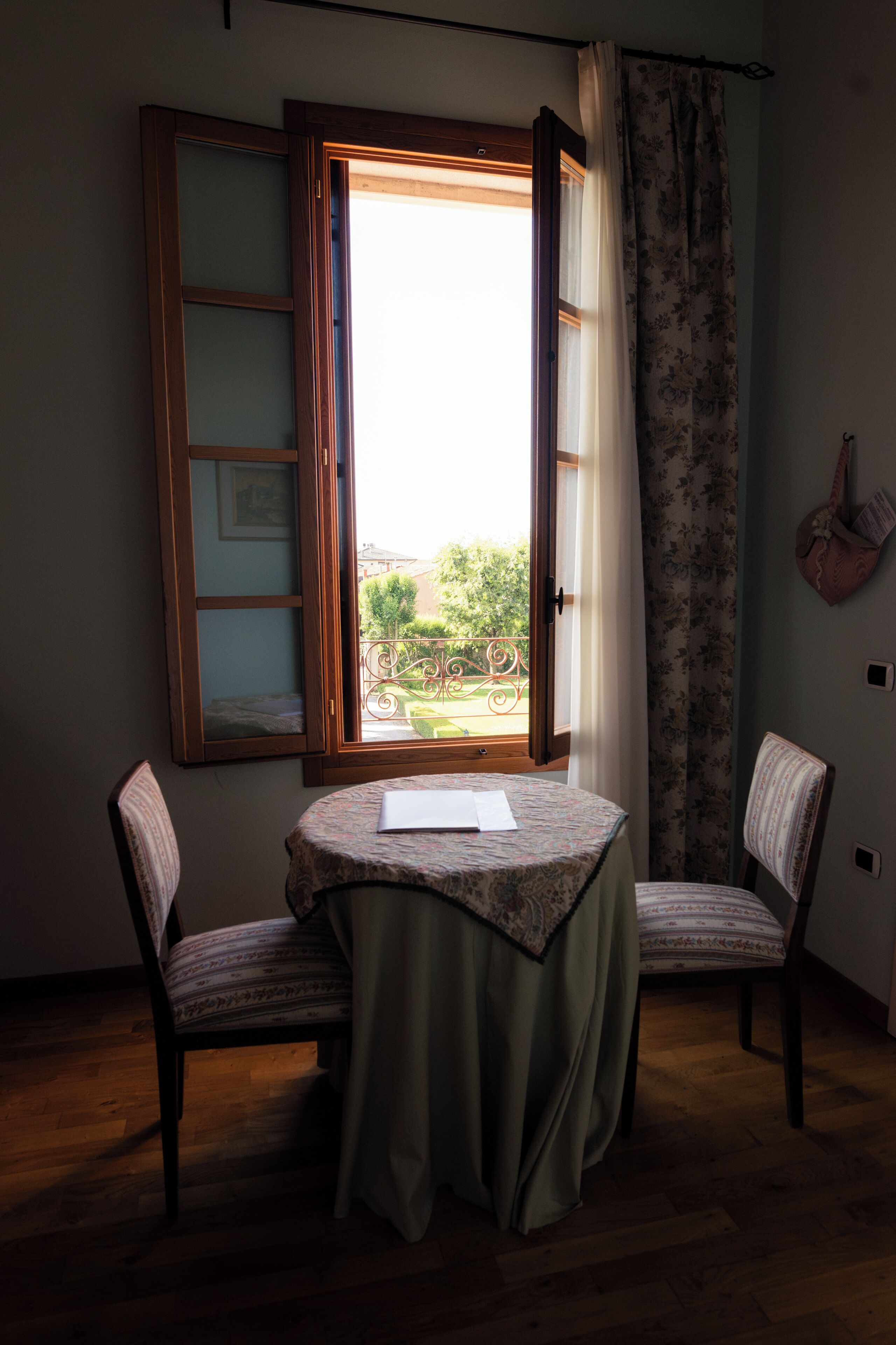 Una habitación con piso de madera, una mesa con mantel estampado, dos sillas, una ventana con cortinas abiertas y vista a un balcón con barandilla.