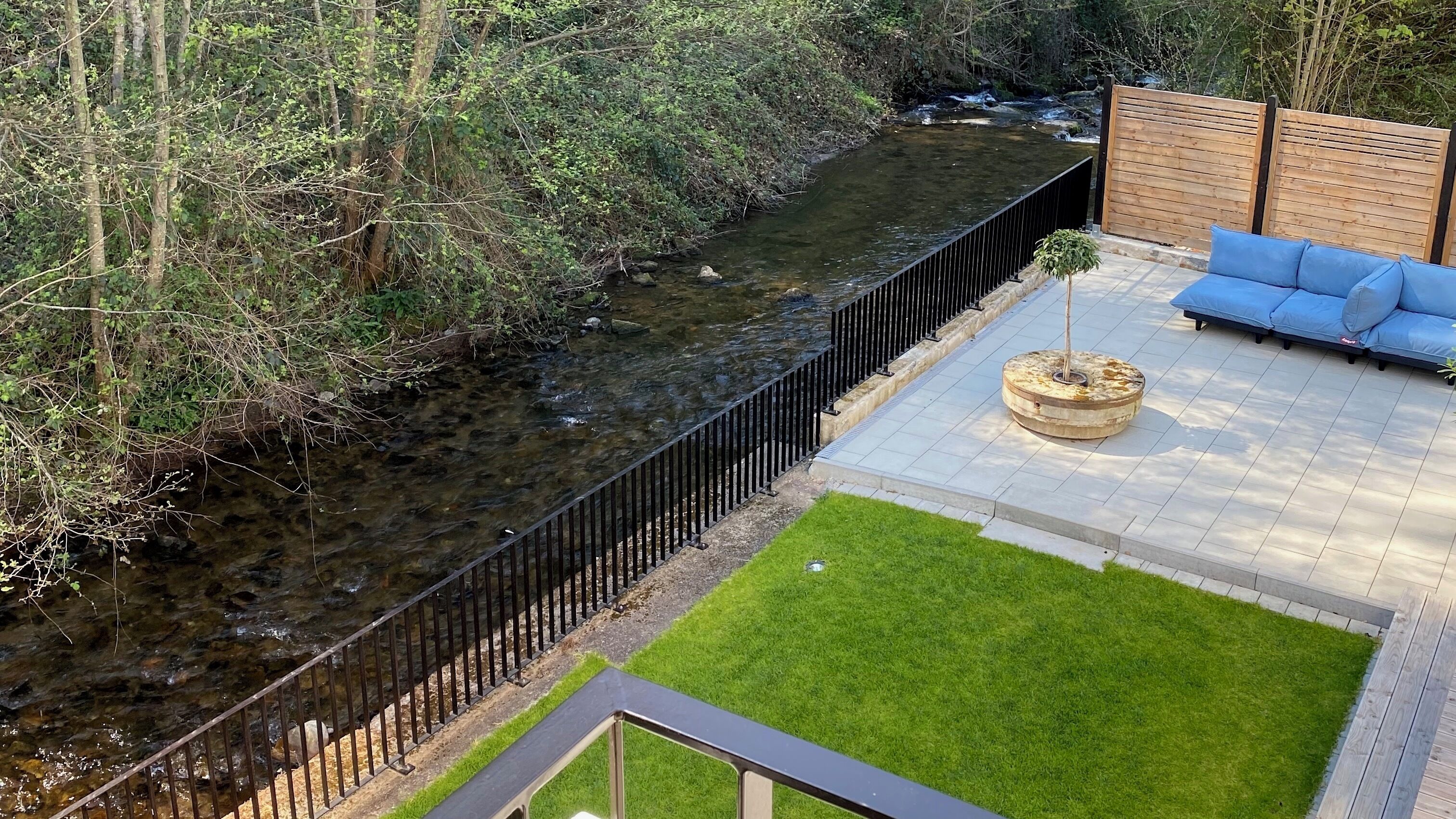 A riverside patio with a wooden fence, a blue sofa, and a small tree planter.