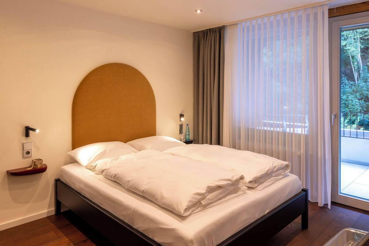 Une chambre d’hôtel moderne avec un grand lit, une tête de lit en bois, une table de chevet avec une lampe et une vue sur l’extérieur à travers des rideaux légers.