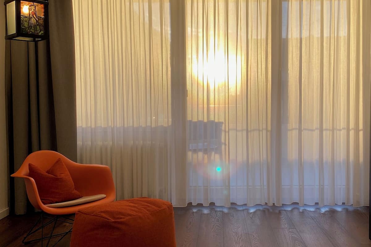 Une pièce avec un lit, une chaise et une grande fenêtre ornée de rideaux légers.
