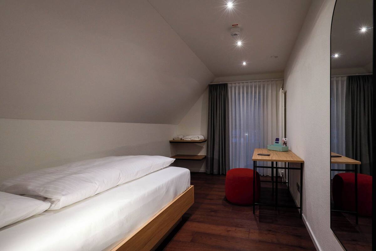 Une chambre d’hôtel moderne avec un lit en bois, un bureau avec une chaise et un petit pouf rouge.