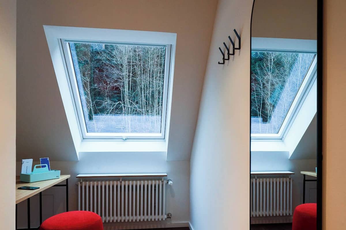 Une chambre moderne avec un plafond en pente, un puits de lumière, un pouf rouge, un grand miroir et un radiateur.