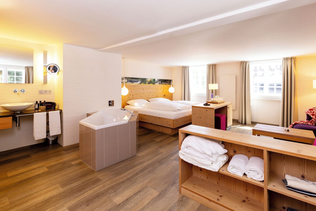 Une chambre d’hôtel avec un plancher en bois, un grand lit, une salle de bain séparée avec une baignoire, une étagère en bois avec des serviettes et une fenêtre avec des rideaux.