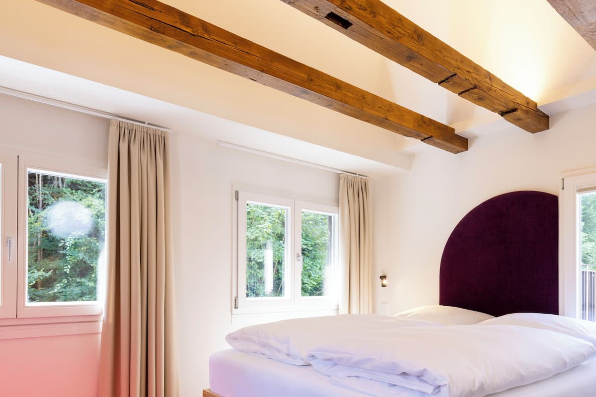 Une chambre avec un plafond en bois, un lit avec une literie blanche, une chaise rouge et des fenêtres avec des rideaux.