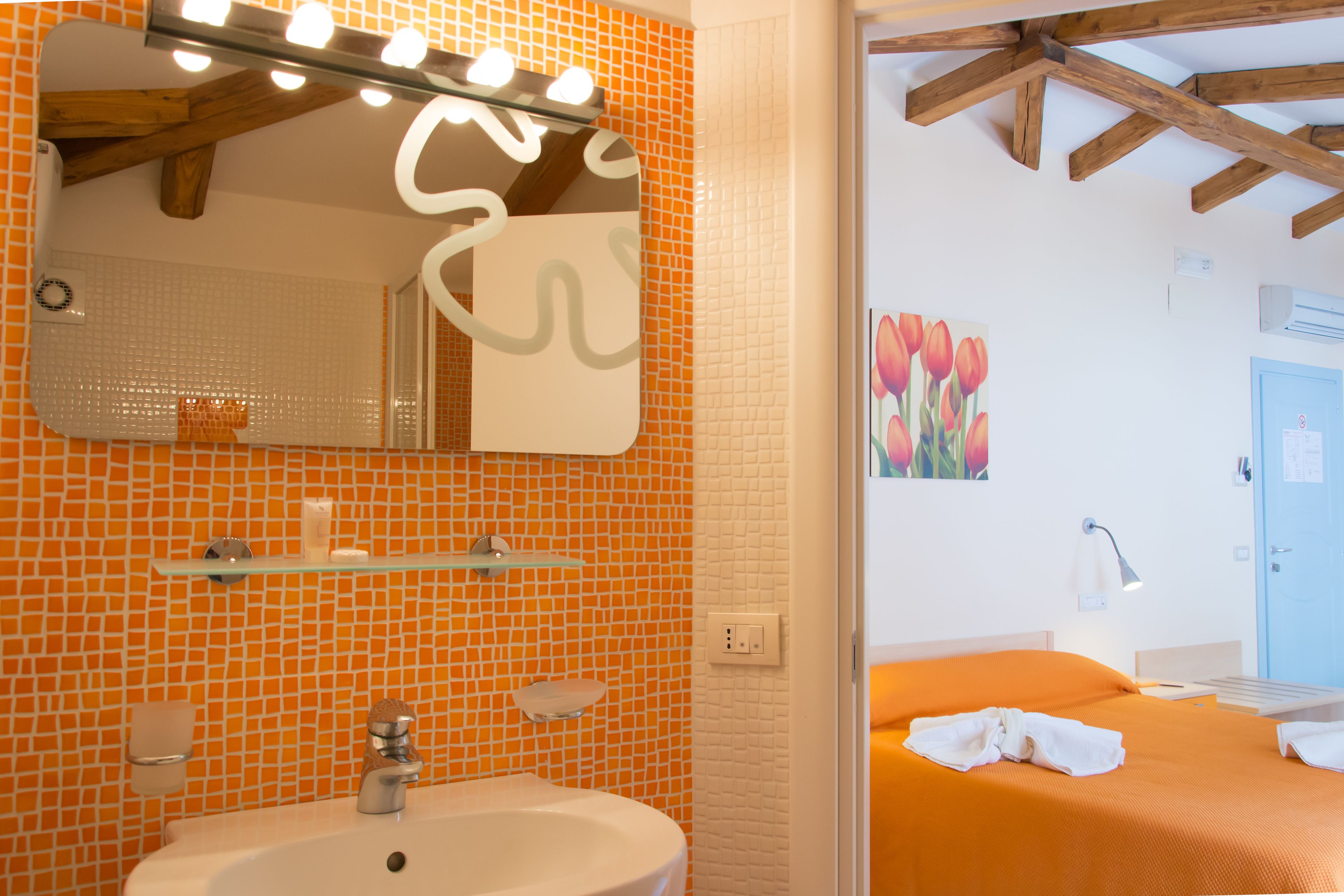 Un baño con pared de mosaicos naranja, un lavabo blanco y un espejo con luces.