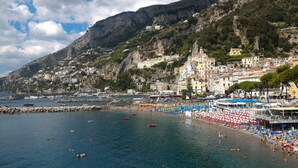 Un pueblo costero con una colina rocosa, una playa con sombrillas y tumbonas, y barcos en el agua.
