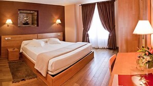 Habitación de hotel con cama de madera, lámparas de noche, una mesita con un jarrón de flores y una ventana con cortinas.