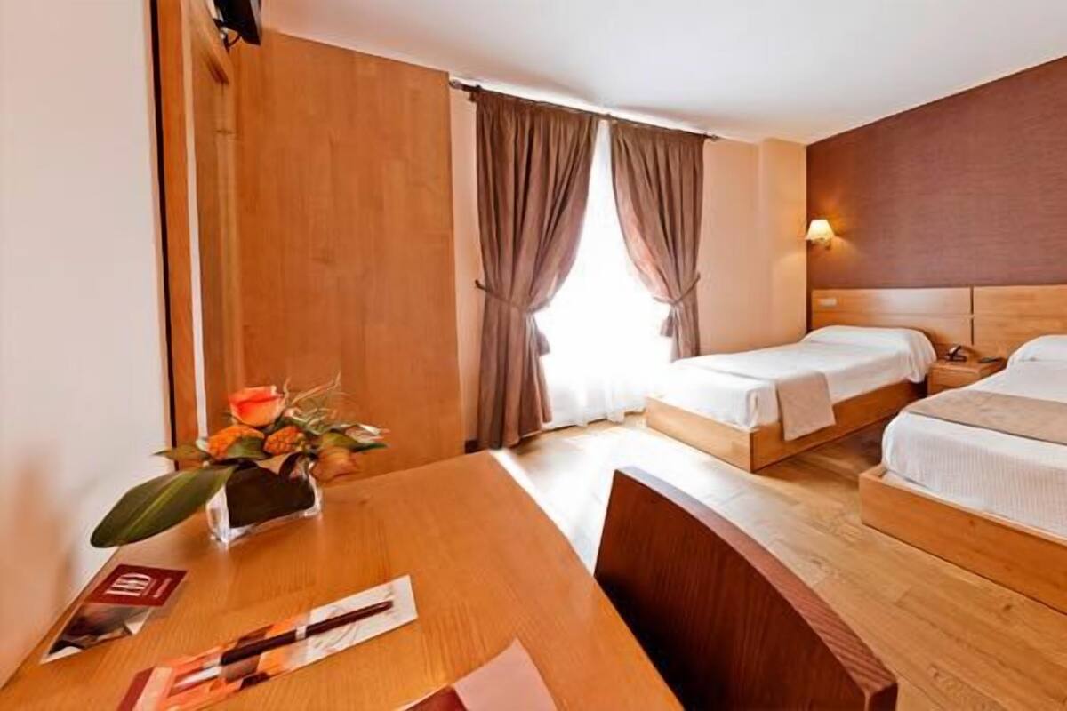 Une chambre d’hôtel avec deux lits simples, un bureau en bois, une chaise, une table ornée d’un vase de fleurs et une fenêtre avec des rideaux.