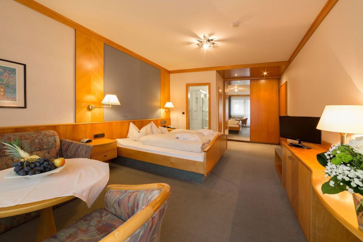 A hotel room with a bed, a desk with a lamp, a chair, a TV, and a bathroom visible in the background.