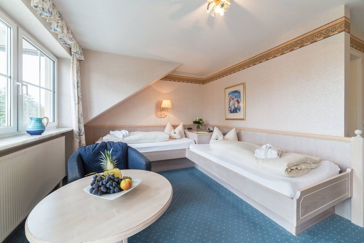 A hotel room with two beds, a dining table with fruit, a lamp, and a painting on the wall.