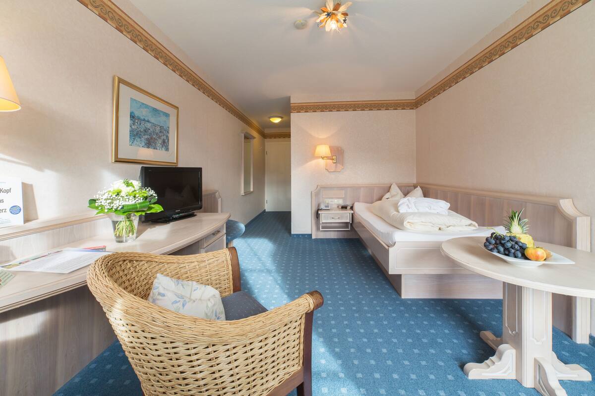 A hotel room with a bed, a desk with a computer, a wicker chair, a small table with fruit, and a wall-mounted TV.