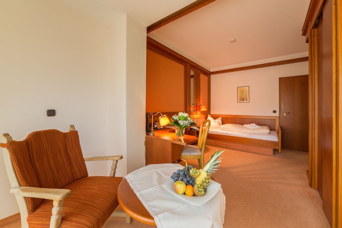 A hotel room with a single bed, a wooden chair, a small table with fruit, a desk with a lamp, and a painting on the wall.