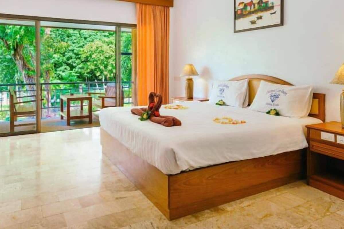 Habitación de hotel con una cama grande, dos lámparas de noche, una mesita de noche de madera, una silla y vistas a un follaje exuberante a través de una puerta corrediza de vidrio.