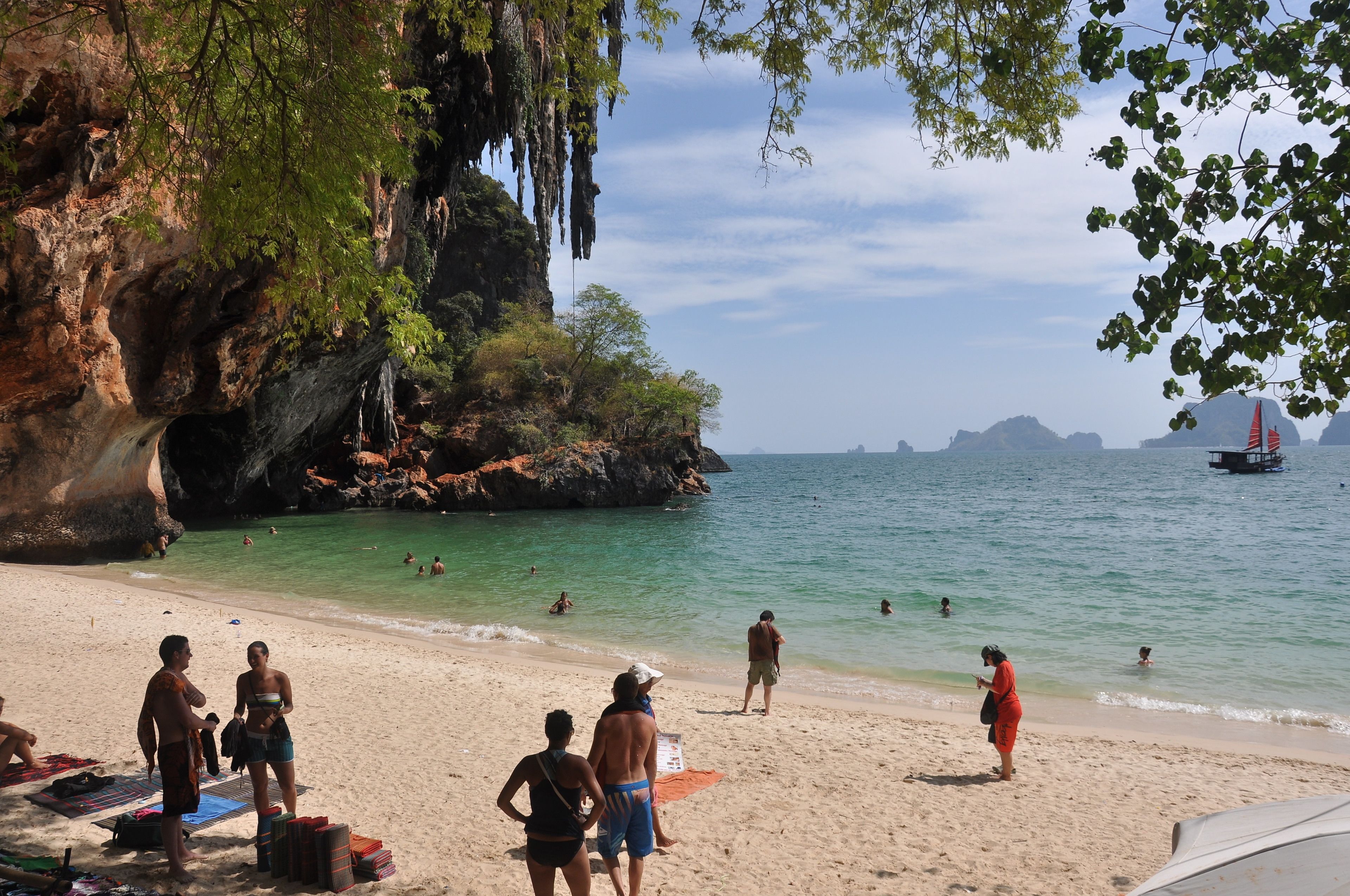 A beach with people swimming and sunbathing, a rocky cliff, and a boat in the distance.