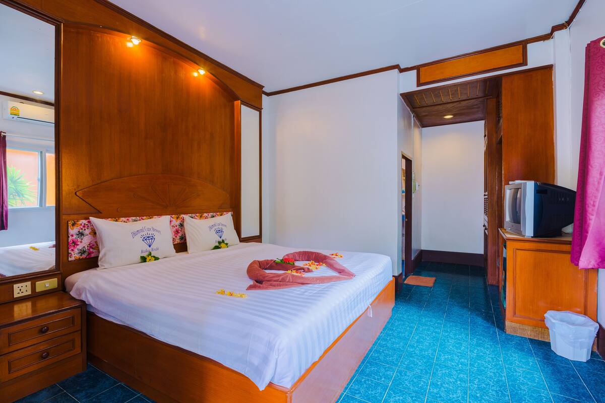 Habitación de hotel con cabecera de madera, cama con sábanas blancas, mesita de noche de madera, televisor sobre un mueble de madera y piso con azulejos azules.