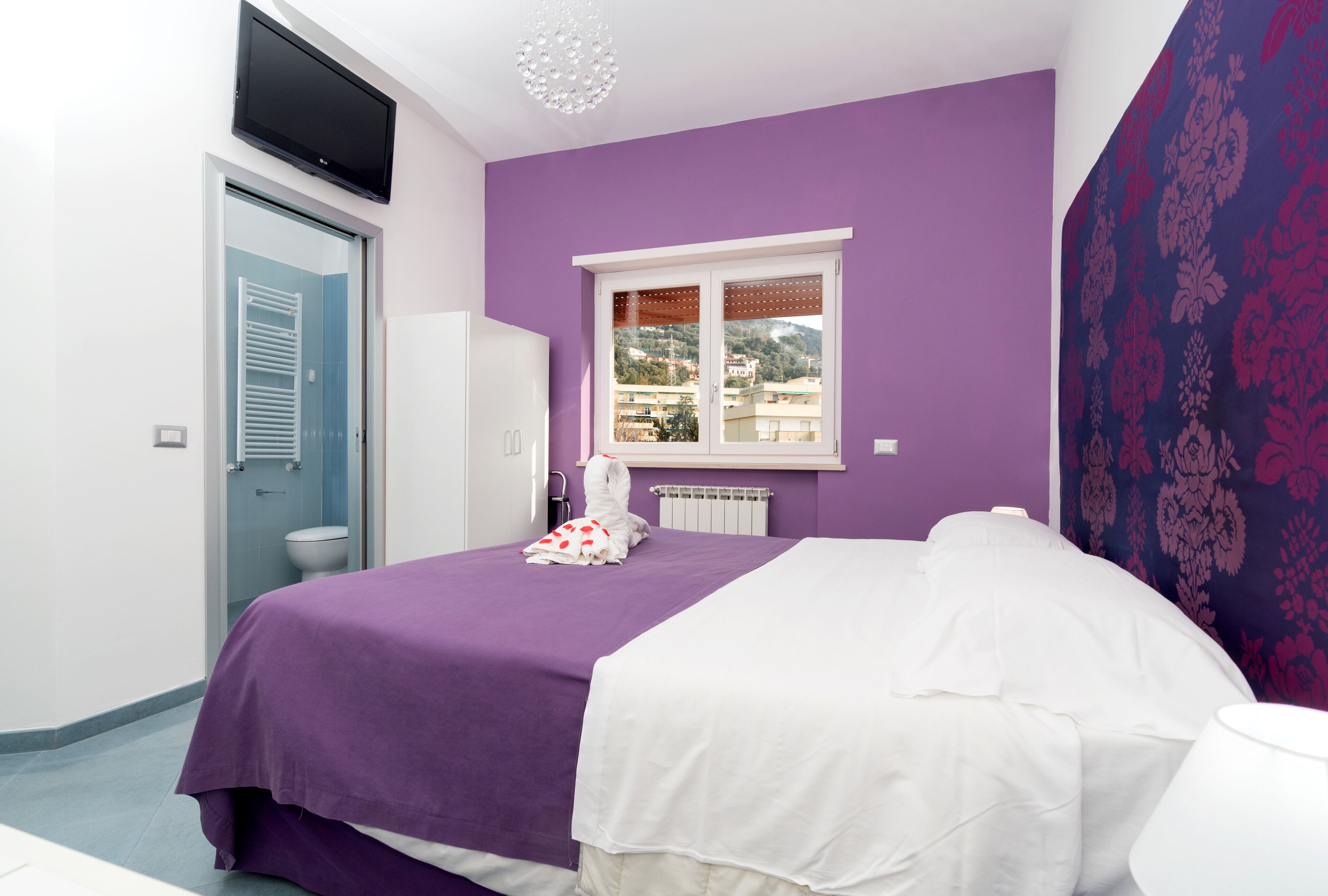 A bedroom with a purple wall, a white bed, and a window with a view of buildings.